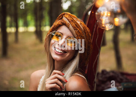 Foto des Blonden reisenden Frau mit stilvollen Accessoires, lächelnd, während in Forest Camp ruhen Stockfoto