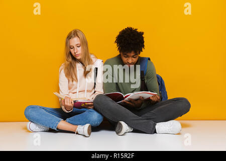 Foto gelangweilt oder verärgert Studenten 16-18 mit Bücher lesen, während sie auf dem Boden mit gekreuzten Beinen über Gelb Hintergrund isoliert Stockfoto