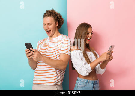 Portrait einer aufgeregt, junges Paar stehen über zwei farbigen Hintergrund, Mobiltelefone zu benutzen, stehen Rücken an Rücken Stockfoto