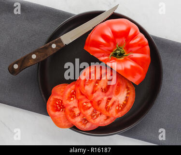 Große, saftige, rote Tomate teil geschnitten auf dunklen Teller mit Messer auf grauer Stoff, Stoff und weißem Marmor Hintergrund Stockfoto