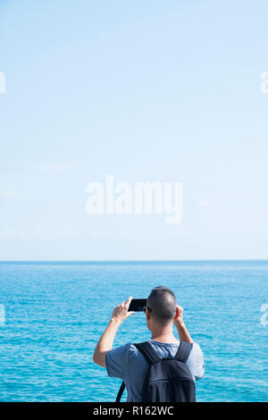 Ein junger weißer Mann, einem blauen T-Shirt tragen und einen Rucksack, von hinten gesehen, ein Bild von dem Meer mit seinen Smartphones, mit einem LARG Stockfoto