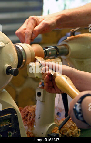 Nahaufnahme der Hand eines älteren männlichen Master woodturner Lehre eine junge weibliche Auszubildende wie eine hölzerne Schüssel auf einer Drehmaschine zu drehen Stockfoto