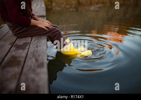 Kind in gelben Gummistiefeln sitzt auf Holzbrücke und legt ihre Beine im Wasser auf dem Hintergrund des Flusses. Nahaufnahme. Stockfoto