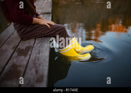 Kind in gelben Gummistiefeln sitzt auf Holzbrücke und legt ihre Beine im Wasser auf dem Hintergrund des Flusses. Nahaufnahme. Stockfoto