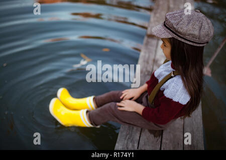 Kind, Mädchen in gelben Gummistiefeln und Kappe sitzt auf Holzbrücke und legt ihre Beine im Wasser auf dem Hintergrund des Flusses. Stockfoto