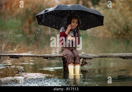 Kind, Mädchen mit Schirm sitzt auf Holzbrücke und lächelt auf dem Hintergrund des Flusses in Regen. Stockfoto