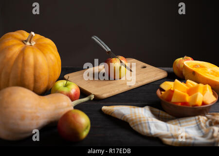 Auf einer hölzernen schwarz Tisch liegen Kürbisse in verschiedenen Größen und Formen, reife Äpfel, ein Geschirrtuch und eine Schüssel von geschnittenen Kürbis. Stockfoto