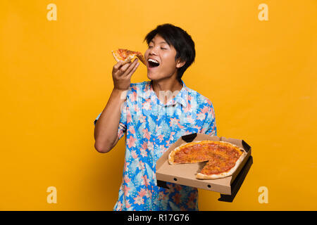 Bild der glücklichen jungen asiatischen Mann stand auf gelben Hintergrund isoliert essen Pizza. Stockfoto