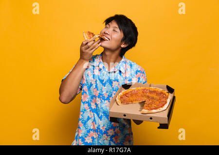 Bild der glücklichen jungen asiatischen Mann stand auf gelben Hintergrund isoliert essen Pizza. Stockfoto