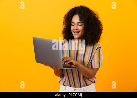 Portrait Of Happy afrikanische Frau stehend über gelb isoliert, mit Laptop Stockfoto