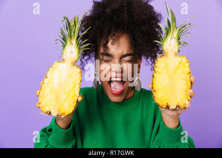 Bild von schreienden schönen emotionalen neugierige junge hübsche afrikanische Frau über violette Hintergrund holding Ananas isoliert. Stockfoto