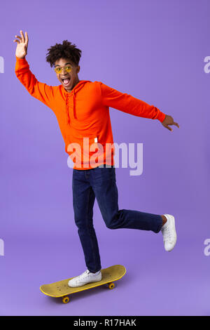 In voller Länge Porträt einer fröhlicher junger afrikanischer Mann reiten auf einem Skateboard über violett Hintergrund isoliert Stockfoto