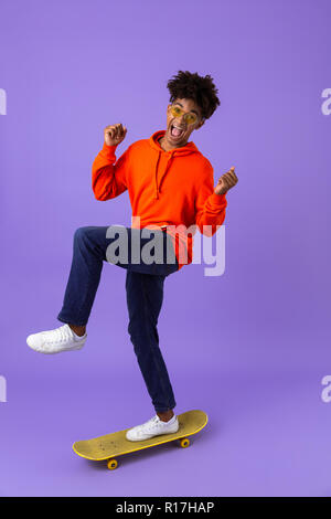 In voller Länge Porträt von einem aufgeregten Jungen afrikanischen Mann reiten auf einem Skateboard über violett Hintergrund isoliert Stockfoto