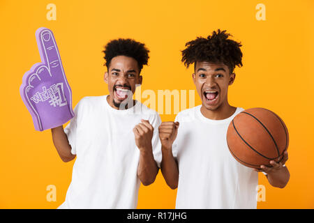 Zwei fröhliche afrikanischer Mann Freunde auf gelben Hintergrund beobachten Basketball Spiel isoliert, Holding foam Handschuh und ein Basketball Stockfoto