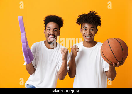 Zwei freudige afrikanischer Mann Freunde auf gelben Hintergrund beobachten Basketball isoliert, Holding foam Handschuh und ein Basketball Stockfoto
