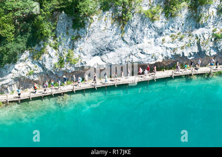 Touristen auf einem Wanderweg zu den Teichen mit türkisfarbenem Wasser und einem hohen Felsen. Blick von oben. Nationalpark Plitvicer Seen, Kroatien Stockfoto