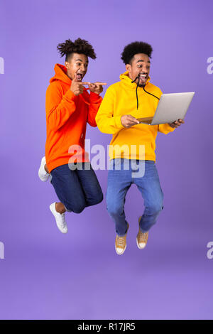 Volle Länge von zwei glücklichen männlichen afrikanischen Freunde in bunten Hoodies über violett Hintergrund isoliert springen, holding Laptop Stockfoto