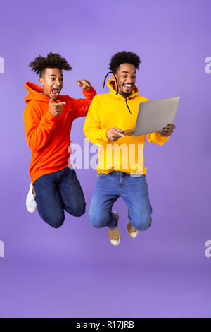 Volle Länge von zwei aufgeregt männlichen afrikanischen Freunde in bunten Hoodies über violett Hintergrund isoliert springen, holding Laptop Stockfoto