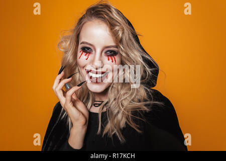 Lächelnde blonde furchtsame Frau Hexe in schwarzen Mantel suchen Kamera über orange isoliert Stockfoto