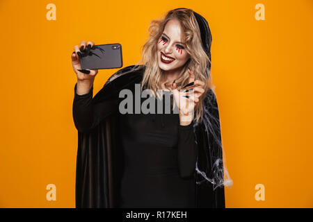 Hübsche Blondine lady Hexe mit unheimlichen Make-up machen selfie auf Smartphone und lächelnd über orange isoliert Stockfoto