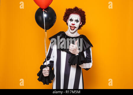 Zornigen Mann in Scary clown Halloween Kostüm holding Ballons über Gelb Hintergrund isoliert gekleidet, mit Daumen hoch Stockfoto