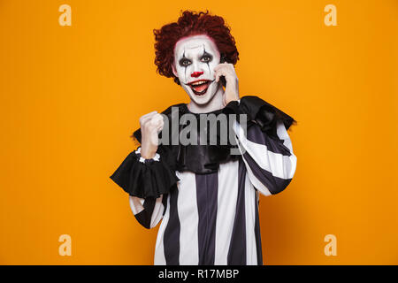 Gerne scary Clown mit Make up Sprechen auf Smartphone und die Gewinner Geste über orange isoliert Stockfoto