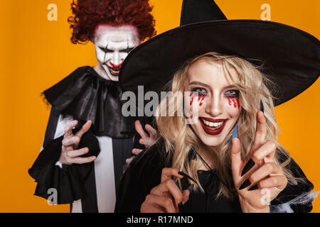 Beängstigend, Zauberer und Clown in Kostümen und mit Halloween Make-up suchen und lächelnd in die Kamera über orange isoliert Stockfoto