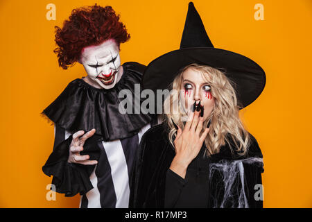 Erschreckend Clown mit Halloween Make-up scary blonde Hexe in schwarzen Hut über orange isoliert Stockfoto