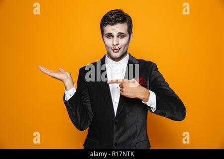 Foto von gruseligen zombie Bräutigam Tragen von klassischen Anzug und Halloween Make-up Holding copyspace auf Palm über Gelb Hintergrund isoliert Stockfoto