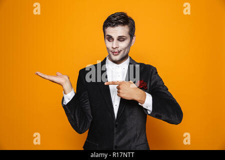 Foto von erschreckend zombie Bräutigam Tragen von klassischen Anzug und Halloween Make-up Holding copyspace auf Palm über Gelb Hintergrund isoliert Stockfoto