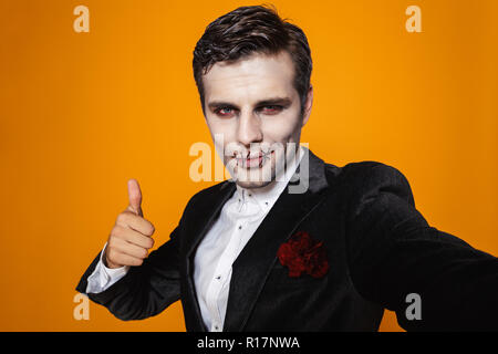 Lächelnd schöner Mann Zombie mit Halloween Make-up, Daumen hoch und selfie über orange isoliert Stockfoto