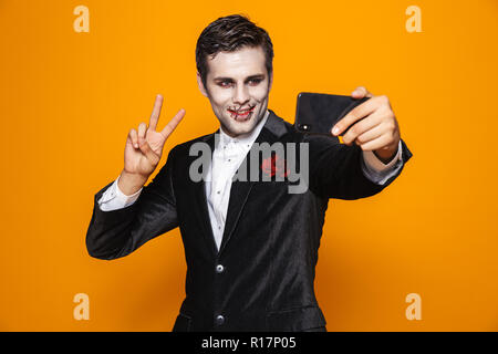 Lächelnd schöner Mann Zombie mit Halloween Make-up zeigt Frieden Geste und selfie auf dem Smartphone über orange isoliert Stockfoto