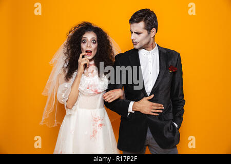 Foto der jungen Zombie paar Bräutigam und Braut mit einem Hochzeitskleid Outfit und Halloween Make-up von der Kamera über Gelb Hintergrund suchen Stockfoto
