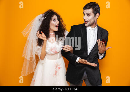 Foto von Happy zombie Paar Bräutigam und Braut mit einem Hochzeitskleid Outfit und Halloween Make-up über Gelb Hintergrund lachen Stockfoto