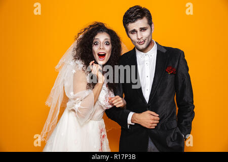 Foto von erschreckend Zombie paar Bräutigam und Braut mit einem Hochzeitskleid Outfit und Halloween Make-up an Suchen Sie isoliert auf gelbem Hintergrund Stockfoto