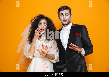 Foto von erschreckend Zombie paar Bräutigam und Braut mit einem Hochzeitskleid Outfit und Halloween makeup erschrecken sie isoliert auf gelbem Hintergrund Stockfoto