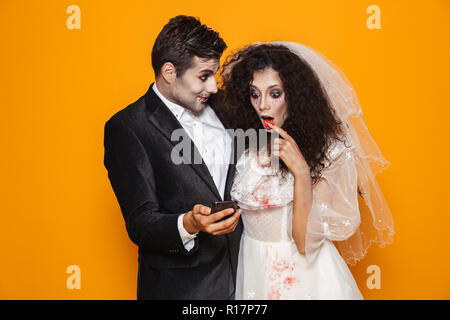 Foto von Angeregten zombie Paar Bräutigam und Braut mit einem Hochzeitskleid Outfit und Halloween Make-up mit Smartphone über Gelb Hintergrund isoliert Stockfoto