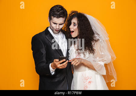 Foto der schönen Zombie paar Bräutigam und Braut mit einem Hochzeitskleid Outfit und Halloween Make-up mit Smartphone über Gelb Hintergrund isoliert Stockfoto