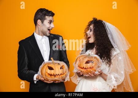Foto der jungen Zombie paar Bräutigam und Braut mit einem Hochzeitskleid Outfit und Halloween Make-up Holding geschnitzten Kürbis über Gelb Hintergrund isoliert Stockfoto