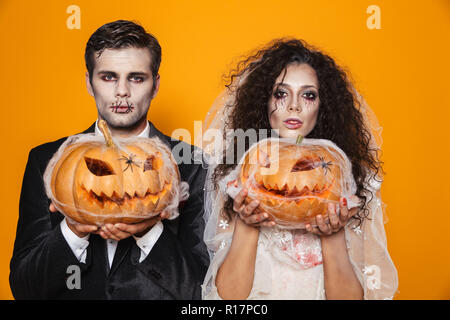 Foto von scary Zombie paar Bräutigam und Braut tragen Outfit und Halloween Make-up Holding geschnitzten Kürbis über Gelb Hintergrund isoliert Stockfoto