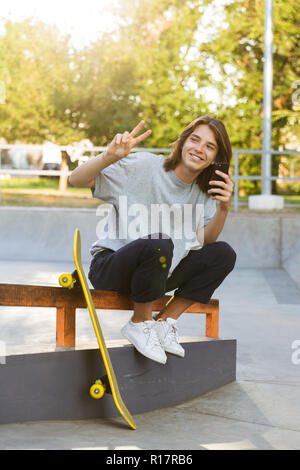 Bild von fröhlichen jungen Skater Kerl sitzen im Park mit über Handy übersicht Frieden Geste Skateboard. Stockfoto
