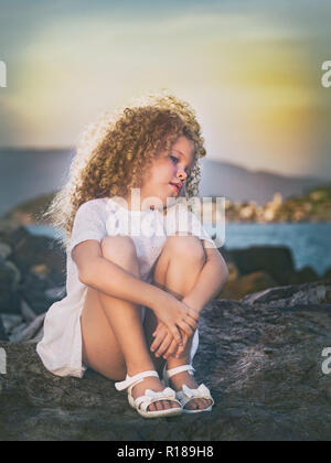 Kleine Mädchen in weißem Kleid auf einem Felsen in der Nähe eines Meeres Stockfoto