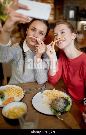 Zwei lustige Mädchen selfie mit Pommes frites zwischen der Nase und Lippen, während Zeit in fast food Cafe Stockfoto