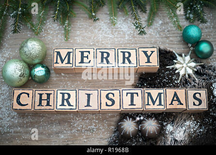 Holzwürfel mit Buchstaben Form der englische Text frohe Weihnachten. Sie liegen auf einem Holz Hintergrund. Tannenzweigen und Weihnachten Kugeln sind. Stockfoto