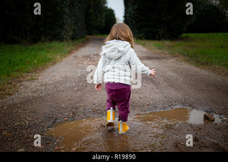 Kleinkind Mädchen, Rückansicht, Planschen in Pfützen in schlammigen Landstrasse mit gelber regen Stiefel. Gegenstand in der Mitte. Stockfoto