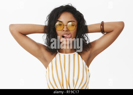 Flirty feminine und elegante zuversichtlich african american Female in modischen Sonnenbrillen und gestreiften Oberteil, Hände hinter dem Kopf in entspannter und Chill darstellen Stockfoto