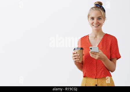 Taille - geschossen von chamring blonde Frau mit gekämmte Haare, die eine neue Tasse Kaffee und Smartphone Lächeln freudig an Kamera in Pause in afternoo Stockfoto