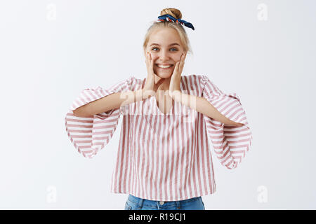 Studio shot zufrieden grinsend optimistische Frau mit blonden Haaren mit Stirnband und gestreifte Bluse quetschen Wangen mit Palmen und breit grinsend her Stockfoto