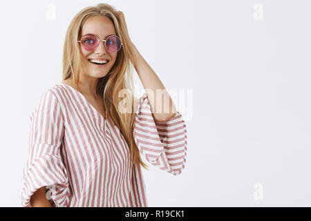 Studio shot von stilvollen und modischen urban fashion Blogger mit runden Sonnenbrille und Rosa gestreifte Bluse, Haar berühren und lächelnd Freude Stockfoto