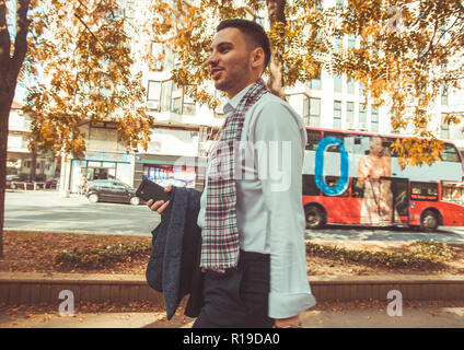 Urban Geschäftsmann, mit einem Lächeln auf seinem Gesicht ist zu Fuß in die Stadt und suchen ernsthafte während seinen Mantel und Telefon in der Hand halten Stockfoto